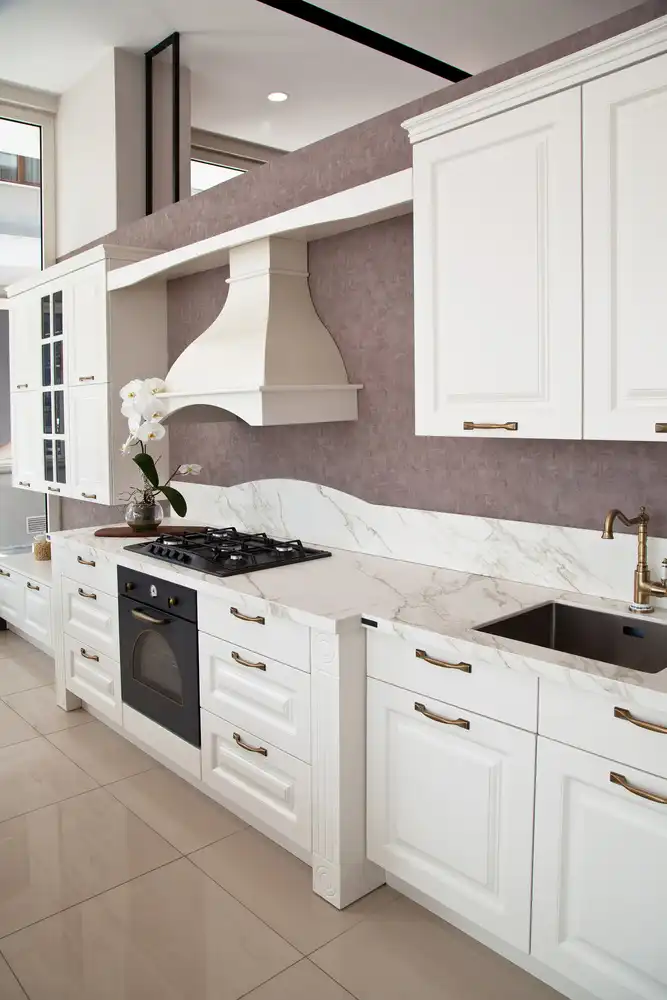 Modern white kitchen cabinet design with marble countertops, gold hardware, stove, sink, and white orchid decoration in natural light