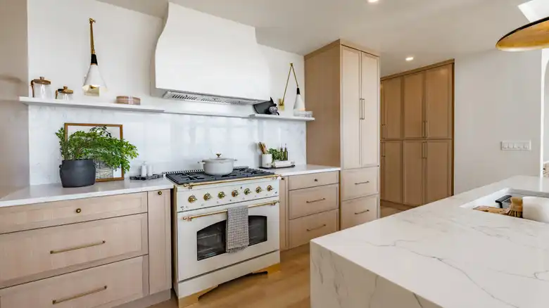 Bright kitchen featuring light natural wood flat-panel cabinets, white marble backsplash and countertops, a large custom white hood, and a retro-style cream and gold range.