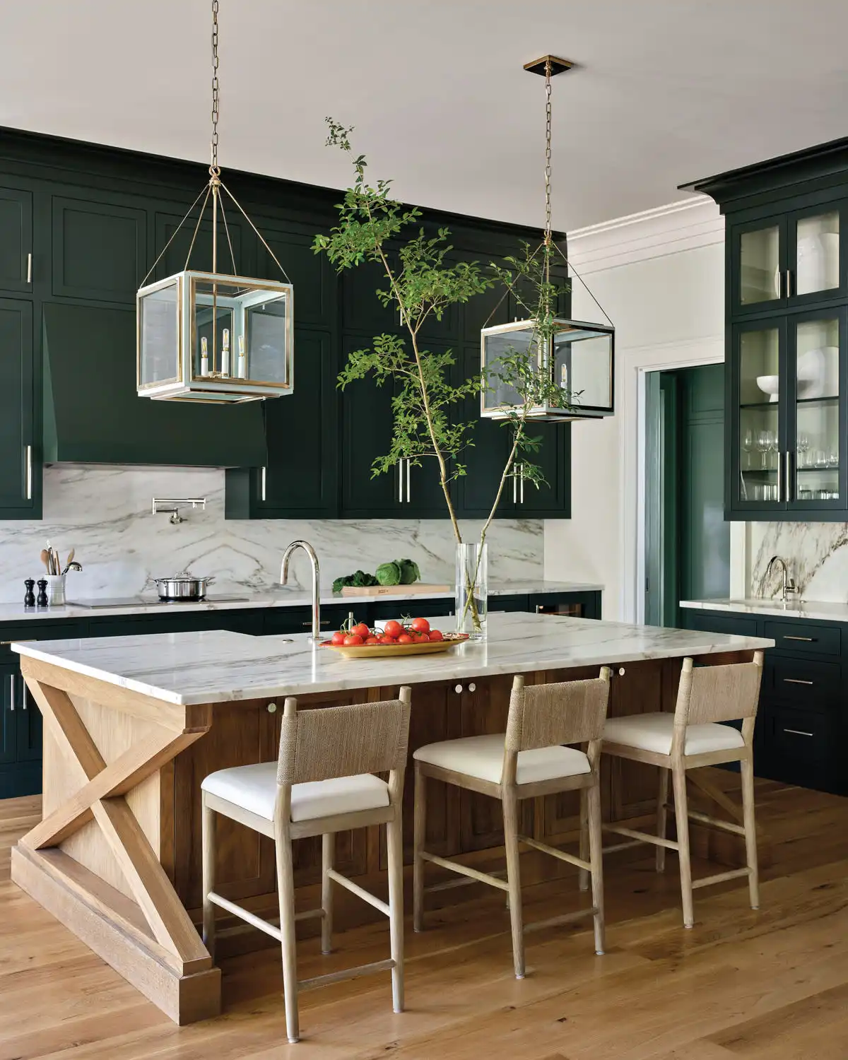 Luxury traditional kitchen featuring dark forest green Shaker cabinets, a large natural wood island with an X-brace base, white marble countertops and backsplash, and brass lantern pendant lights.