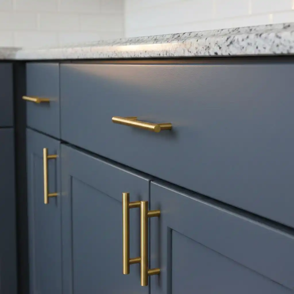 A close-up view of modern kitchen cabinets painted in a dark, dusty blue or navy gray color, fitted with brushed gold (brass) bar pull handles. The top drawer features a long horizontal pull, while the lower cabinets have two stacked vertical pulls. The countertop is speckled granite, and the backsplash is white subway tile. Focus keyword: choose the best gold cabinet hardware design.