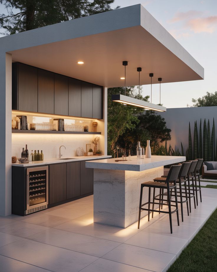 A contemporary backyard outdoor kitchen showcasing minimalist design with dark flat-panel cabinetry and a striking marble-effect waterfall island with integrated under-counter lighting. Four dark high-back woven bar stools are seated at the counter, which features a long minimalist black linear pendant light.